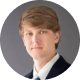Headshot of a young man with side-swept light brown hair wearing a dark suit and tie, set against a neutral gray background. He has a serious, confident expression.