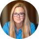 Tonya Weinzettle A woman with long blonde hair and glasses, wearing a blue shirt, sits indoors in front of a blurred background, looking directly at the camera with a slight smile.